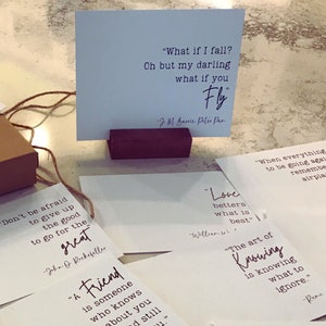May include: A collection of inspirational quote cards with white backgrounds and black text. The cards feature quotes from authors such as J.M. Barrie, John D. Rockefeller, and Rumi. The cards are displayed on a wooden stand.