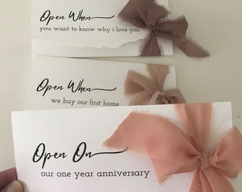 May include: Three white cards with black text and pink ribbon bows. The text on the cards reads "Open When you want to know why I love you", "Open When we buy our first home", and "Open On our one year anniversary".