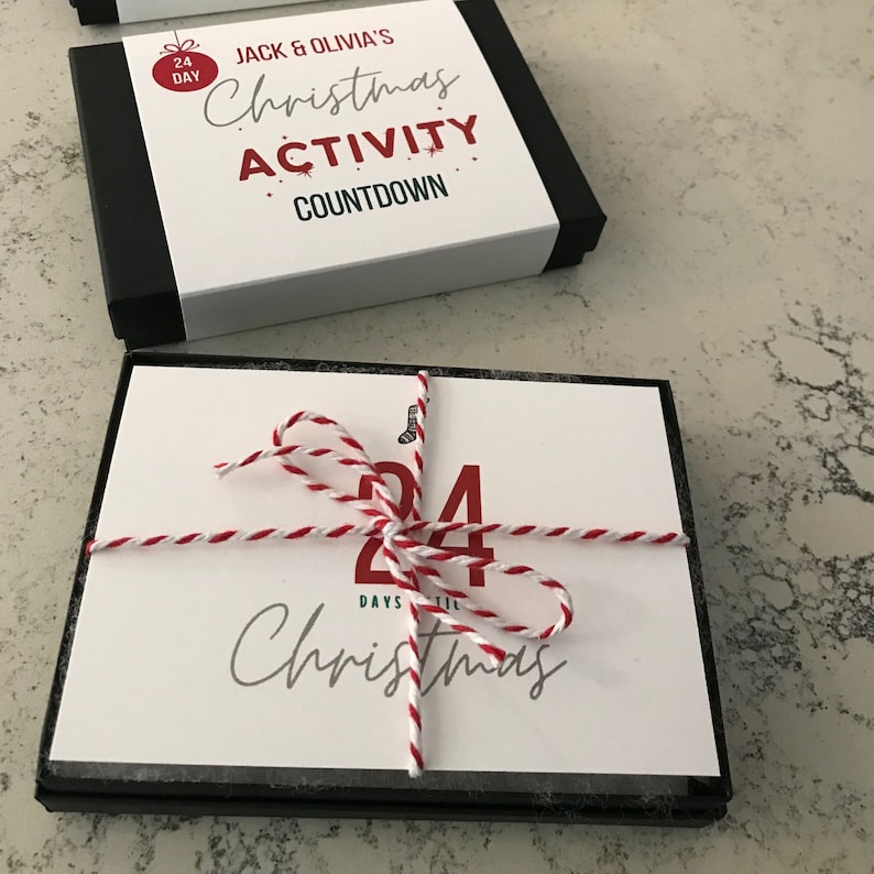 Puede incluir: Una caja blanca con ribete negro, atada con una cuerda roja y blanca. La caja tiene una etiqueta blanca con el texto "24 Days til Christmas" y un n&uacute;mero rojo "24". La caja est&aacute; etiquetada "Jack & Olivia's Christmas Activity Countdown".