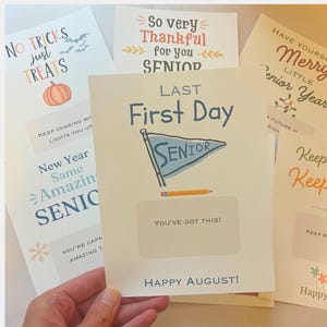 May include: A collection of white rectangular cards featuring various seasonal and school-themed graphics and text. One card prominently displays the text 'LAST First Day' above a blue pennant flag labelled 'SENIOR', a pencil, and 'HAPPY AUGUST!'