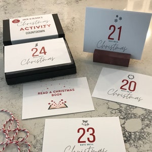 Puede incluir: Un conjunto de actividades de cuenta atr&aacute;s para Navidad con una caja negra y tarjetas blancas. Las tarjetas presentan n&uacute;meros del 20 al 24, las palabras "D&iacute;as hasta Navidad" e ilustraciones festivas. La caja tiene el texto "Jack & Olivia's Christmas Activity Countdown".