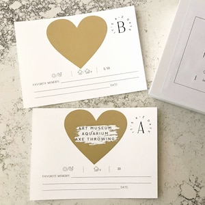 May include: Two white cards with gold hearts. The hearts have text inside. The top card has the letter "B" inside the heart. The bottom card has the text "Art Museum Aquarium Axe Throwing" inside the heart. Both cards have a space for a favorite memory and a date.