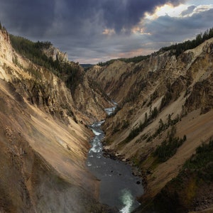 May include: A dramatic view of a canyon in Yellowstone National Park, with a river winding through the centre. The canyon walls are a mix of brown, tan, and grey rock, with some green vegetation growing on the slopes. The sky is a dark grey with a few rays of sunlight breaking through the clouds.