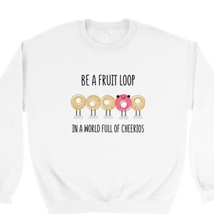 Puede incluir: Sudadera blanca con el texto "BE A FRUIT LOOP IN A WORLD FULL OF CHEERIOS". El texto está en negro. Hay cinco bucles de cereales de dibujos animados, cuatro son beige y uno es rosa. El bucle rosa tiene una cara feliz.