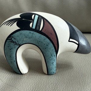 以下が含まれることがあります： クリーム色のボディと塗装されたディテールが特徴のセラミック製のクマの置物。クマは黒、ティール、茶色の幾何学模様が特徴です。「Sandien」というアーティストのサインが見えます。クマは脚を広げた、湾曲した姿勢をしています。