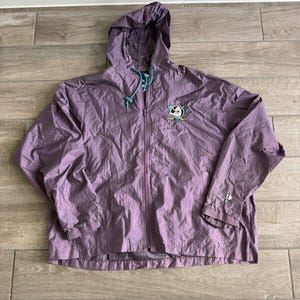 Op de afbeelding: Een paarse jas met capuchon en een volledige ritssluiting. De jas heeft een turquoise trekkoord en het logo van het Anaheim Ducks hockeyteam op de borst. De jas heeft een lichte, gekreukelde textuur.