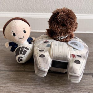 Op de afbeelding: Een pluche speelgoedset met een Millennium Falcon, een Chewbacca-pop en een Han Solo-pop. De Millennium Falcon is lichtgrijs met bedrukte details. Chewbacca is bruin en Han Solo draagt een witte outfit met een blauw vest.
