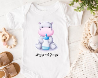 May include: White baby onesie with a cartoon illustration of a gray hippopotamus holding a blue and pink baby bottle. The text "Hungry and Grumpy" is printed below the hippo.