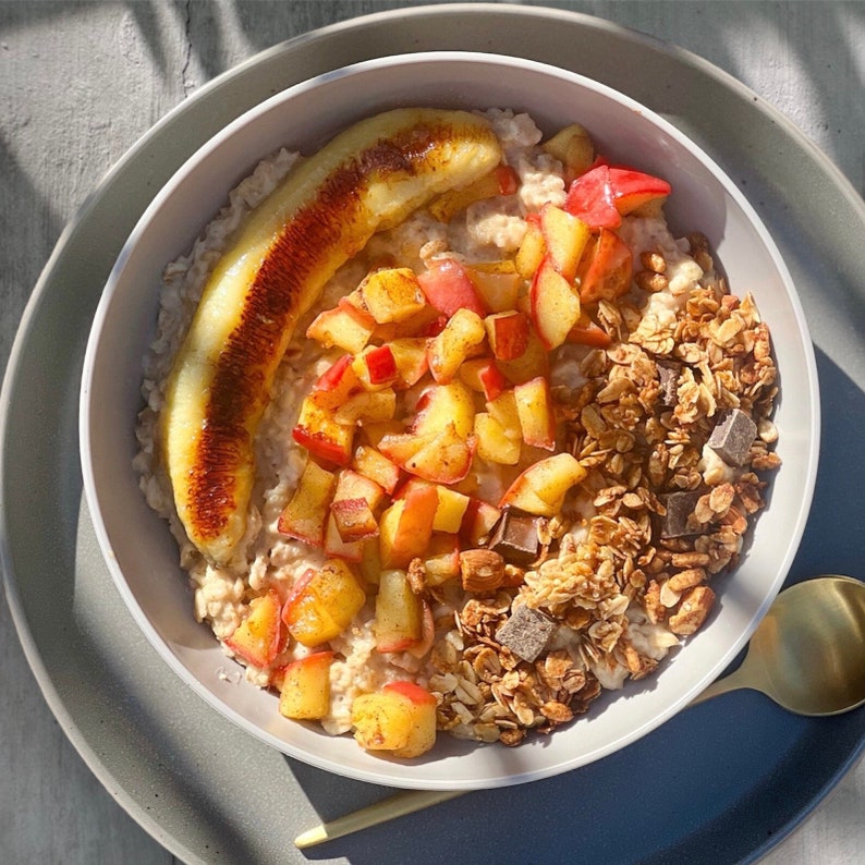 Puede incluir: Un cuenco de avena con pl&aacute;tano caramelizado, melocotones en dados, granola y trozos de chocolate. El cuenco est&aacute; sobre un plato gris, con una cuchara dorada.