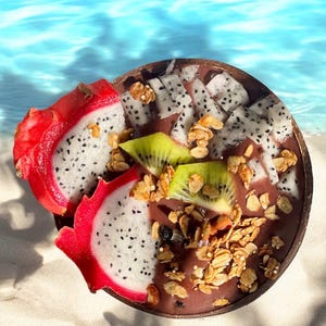 May include: A close-up of a smoothie bowl with dragon fruit, kiwi, and granola. The bowl is made of a natural material and is sitting on a white sandy beach.