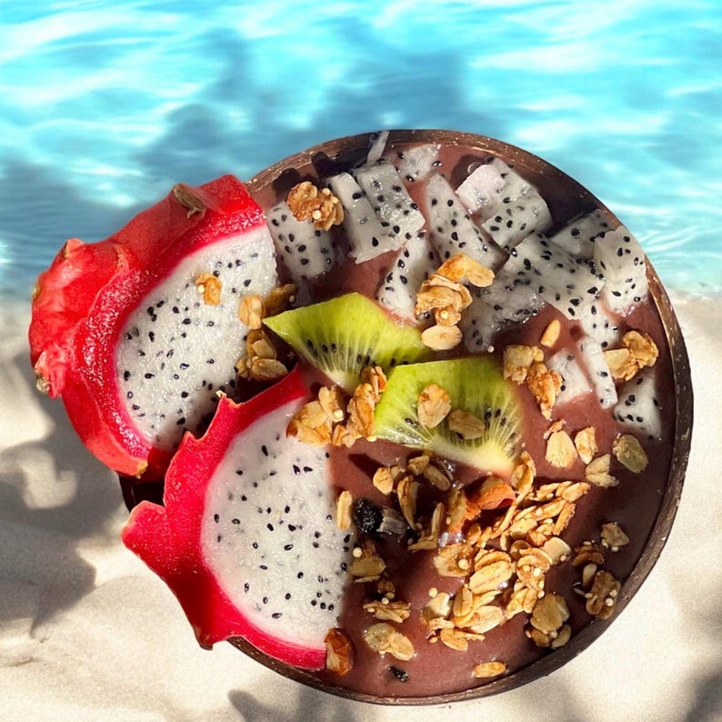 May include: A close-up of a smoothie bowl with dragon fruit, kiwi, and granola. The bowl is made of a natural material and is sitting on a white sandy beach.
