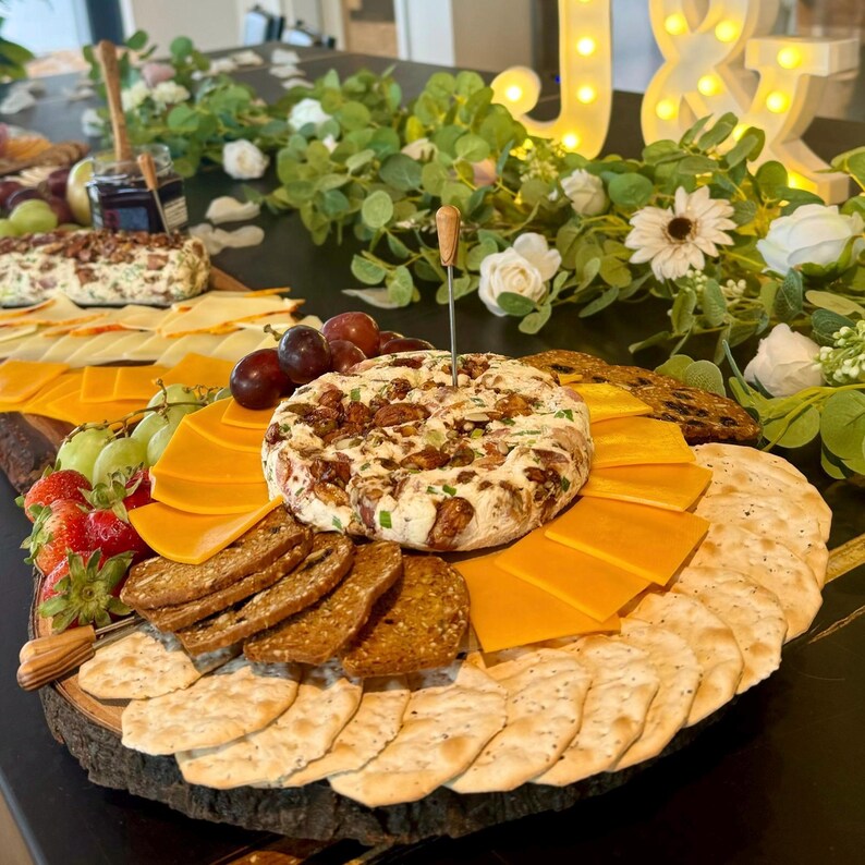 May include: A wooden charcuterie board with a selection of snacks, including crackers, cheese, fruit, and a large round cream cheese spread topped with pecans and spring onions.