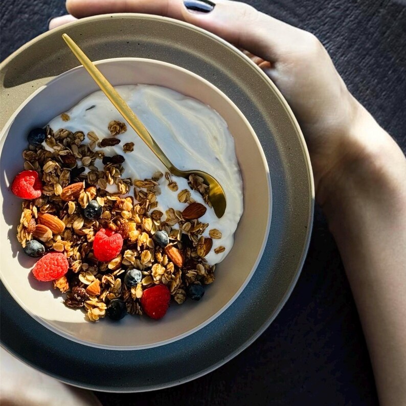 May include: A bowl of granola with yogurt, blueberries, and raspberries. The granola is topped with almonds and a golden spoon is resting on the side of the bowl.
