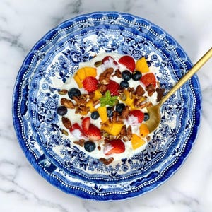K&ouml;nnte beinhalten: Eine blaue und wei&szlig;e Sch&uuml;ssel mit Muster, gef&uuml;llt mit Joghurt, Obst und M&uuml;sli. Die Sch&uuml;ssel steht auf einer wei&szlig;en Marmorfl&auml;che.
