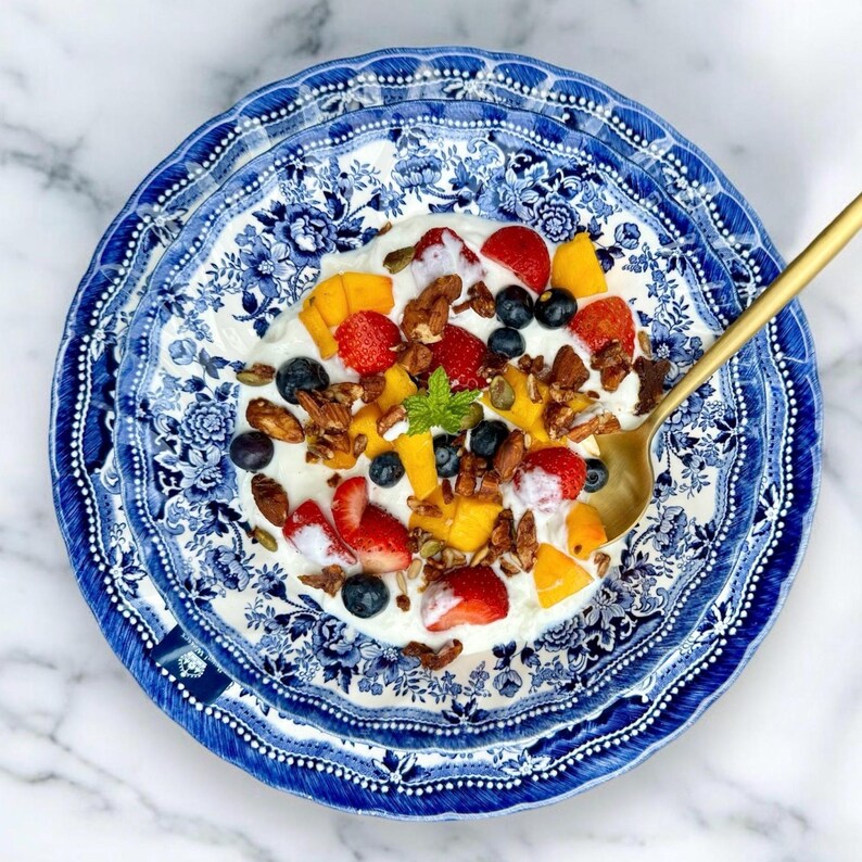 K&ouml;nnte beinhalten: Eine blaue und wei&szlig;e Sch&uuml;ssel mit Muster, gef&uuml;llt mit Joghurt, Obst und M&uuml;sli. Die Sch&uuml;ssel steht auf einer wei&szlig;en Marmorfl&auml;che.