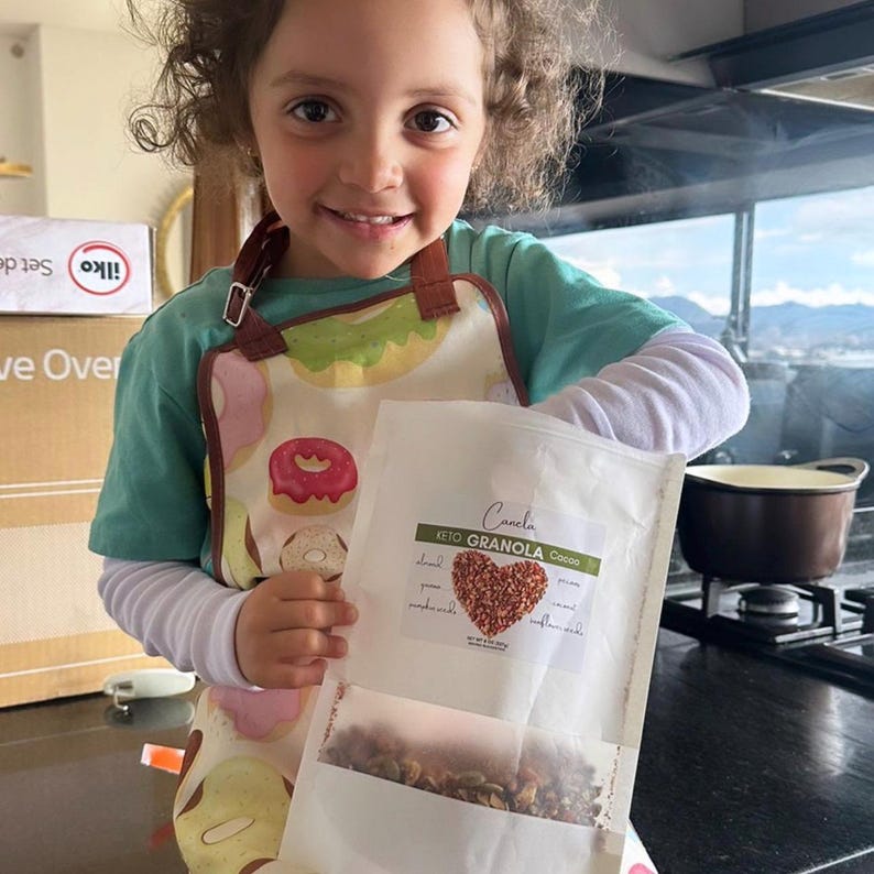 K&ouml;nnte beinhalten: Ein junges Kind tr&auml;gt eine bunte Sch&uuml;rze und h&auml;lt eine T&uuml;te Granola mit dem Text "Canela Keto Granola Coco" auf der Vorderseite. Die T&uuml;te ist transparent und zeigt das Granola im Inneren.