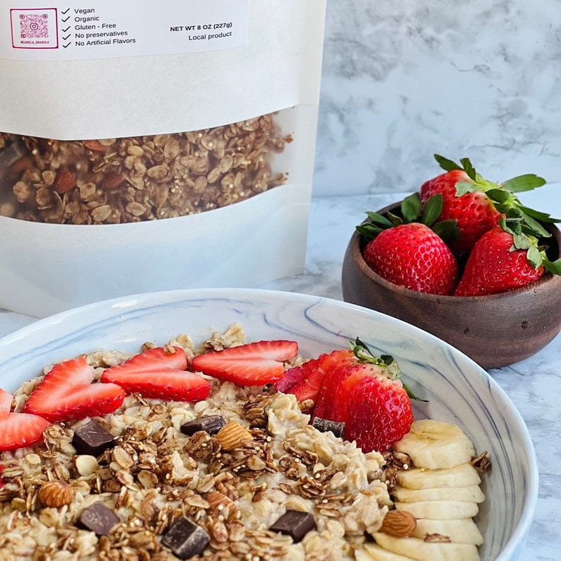 May include: A bowl of oatmeal topped with sliced bananas, strawberries, chocolate chips, almonds, and granola. The oatmeal is a light brown color and the granola is a darker brown color. The strawberries are red and the bananas are yellow. The chocolate chips are dark brown and the almonds are light brown. The bowl is white with a marble pattern. The text on the bag reads: Vegan, Organic, Gluten-Free, No Preservatives, No Artificial Flavors, NET WT 8 OZ (227g), Local product.