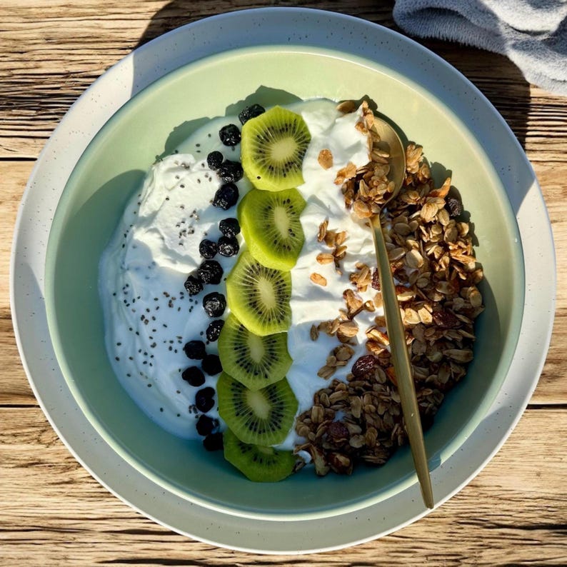Puede incluir: Un cuenco verde claro lleno de yogur, rodajas de kiwi, ar&aacute;ndanos, semillas de ch&iacute;a y granola. Una cuchara dorada descansa en la granola. El cuenco est&aacute; sobre un plato blanco, todo sobre una superficie de madera.