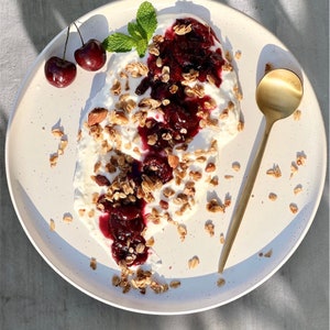 Puede incluir: Un plato de yogur cubierto con granola, compota de cerezas y menta fresca. Dos cerezas est&aacute;n en el lateral del plato. Una cuchara dorada descansa en el plato. El plato es blanco con un dise&ntilde;o moteado.