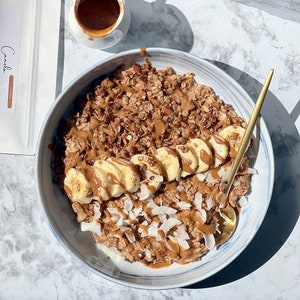 Puede incluir: Un taz&oacute;n de avena cubierto con rodajas de pl&aacute;tano, granola, coco rallado y mantequilla de nueces. Una cuchara dorada descansa en el taz&oacute;n. Una peque&ntilde;a taza blanca de caf&eacute; y una bolsa de "Canela Coffee" est&aacute;n en el fondo.