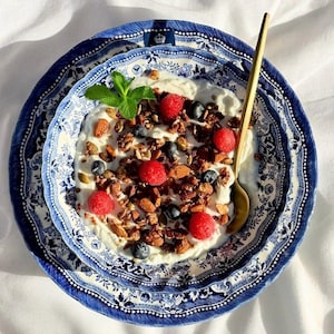 May include: A bowl of yogurt topped with granola, raspberries, and blueberries, served in a blue and white patterned bowl with a gold spoon.
