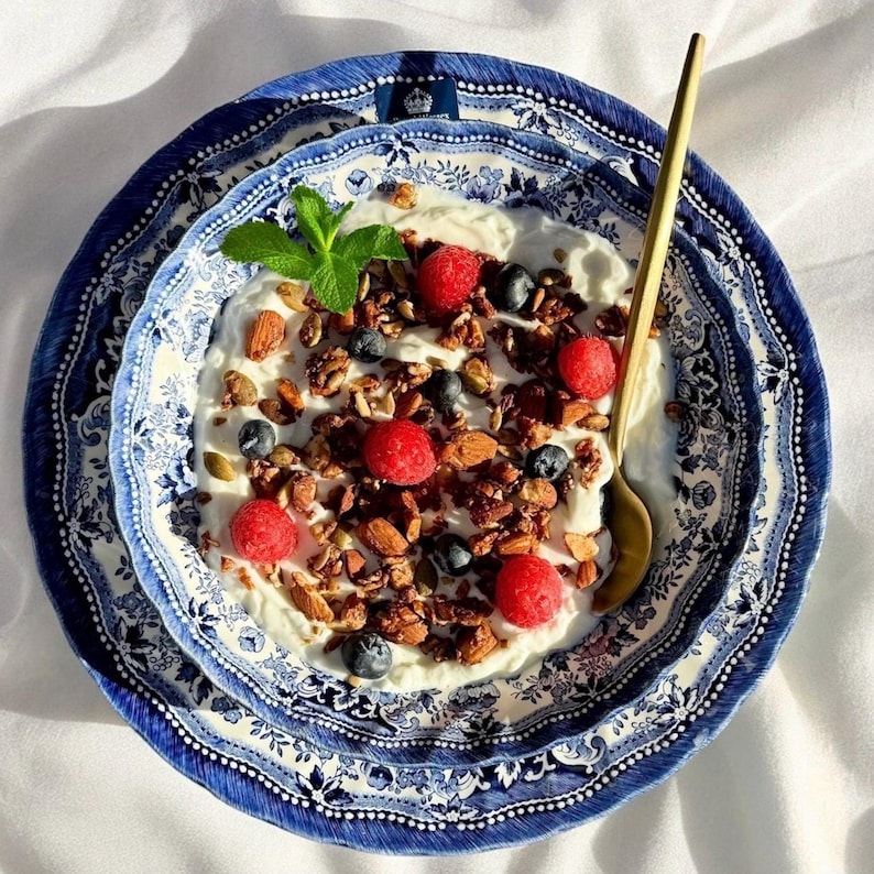 May include: A bowl of yogurt topped with granola, raspberries, and blueberries, served in a blue and white patterned bowl with a gold spoon.