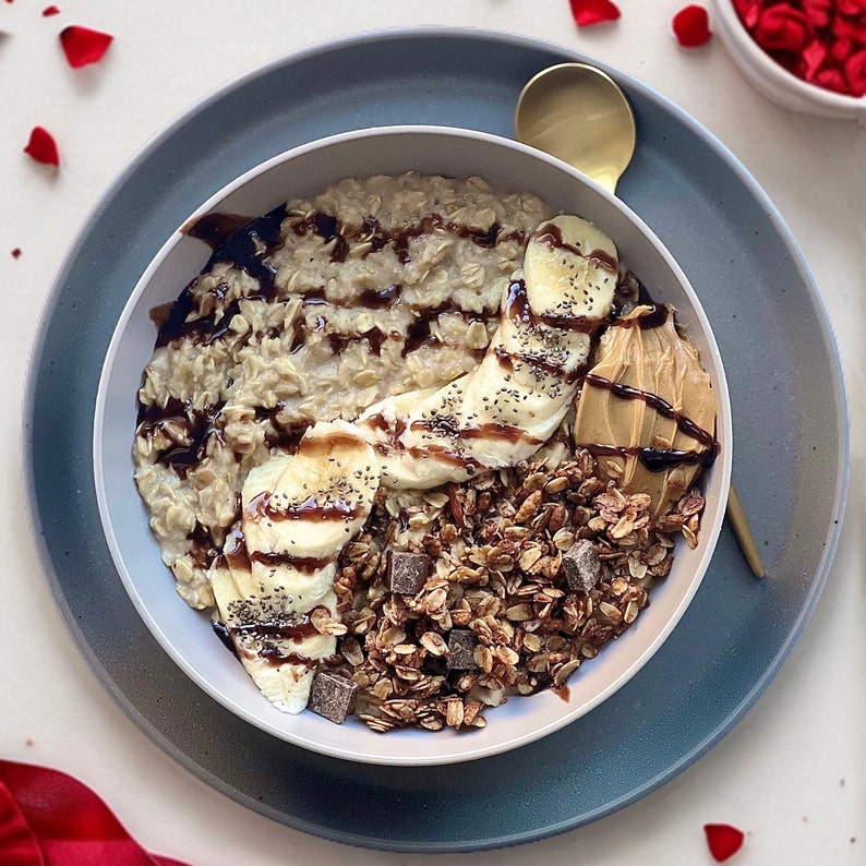 May include: A bowl of oatmeal topped with sliced bananas, chocolate chips, granola, and peanut butter. The oatmeal is drizzled with chocolate sauce.