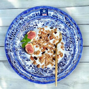 May include: A white bowl containing yogurt, granola, and figs, served on a blue and white patterned plate. The plate features a blue and white floral design with a gold rim. The plate is placed on a white wooden surface.