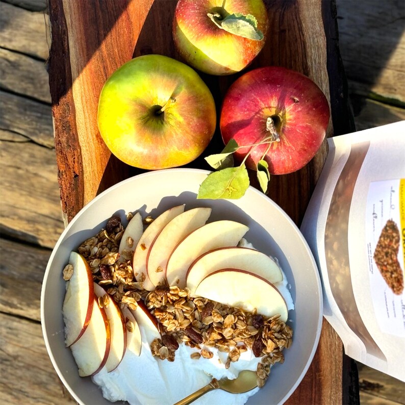 May include: A bowl of yogurt topped with granola, sliced apples, and a few pecans. Three red apples are in the background.