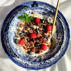 K&ouml;nnte beinhalten: Eine blaue und wei&szlig;e Keramik Sch&uuml;ssel gef&uuml;llt mit Joghurt, Granola, Himbeeren und Blaubeeren. Ein goldener L&ouml;ffel liegt am Rand der Sch&uuml;ssel.