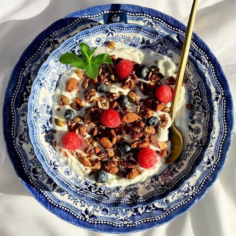 K&ouml;nnte beinhalten: Eine blaue und wei&szlig;e Keramik Sch&uuml;ssel gef&uuml;llt mit Joghurt, Granola, Himbeeren und Blaubeeren. Ein goldener L&ouml;ffel liegt am Rand der Sch&uuml;ssel.