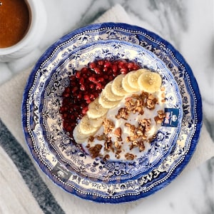 May include: A blue and white patterned bowl filled with yogurt, granola, sliced bananas, and pomegranate seeds.