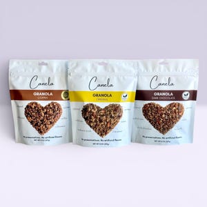 Puede incluir: Tres bolsas blancas de granola "Canela", cada una con un dise&ntilde;o de granola en forma de coraz&oacute;n. Los sabores son Caf&eacute;, Original y Chocolate Negro. Las bolsas indican "Sin conservantes. Sin sabores artificiales."