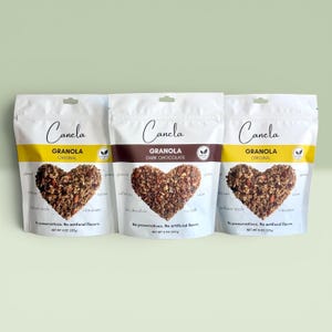 Puede incluir: Tres bolsas blancas de granola Canela, cada una con un grupo de granola en forma de coraz&oacute;n. Las bolsas est&aacute;n etiquetadas como "Original" y "Dark Chocolate". Cada bolsa pesa 227g.