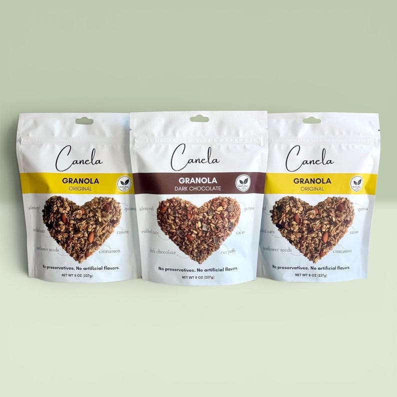 Puede incluir: Tres bolsas blancas de granola Canela, cada una con un grupo de granola en forma de coraz&oacute;n. Las bolsas est&aacute;n etiquetadas como "Original" y "Dark Chocolate". Cada bolsa pesa 227g.