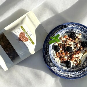 K&ouml;nnte beinhalten: Eine wei&szlig;e Sch&uuml;ssel mit blau-wei&szlig;em Muster, gef&uuml;llt mit Joghurt, Granola, Brombeeren und Mandeln. Die Sch&uuml;ssel steht auf einer wei&szlig;en Oberfl&auml;che. Im Hintergrund befindet sich eine T&uuml;te Keto-Granola.