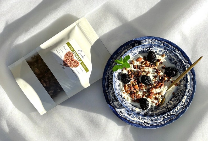 K&ouml;nnte beinhalten: Eine wei&szlig;e Sch&uuml;ssel mit blau-wei&szlig;em Muster, gef&uuml;llt mit Joghurt, Granola, Brombeeren und Mandeln. Die Sch&uuml;ssel steht auf einer wei&szlig;en Oberfl&auml;che. Im Hintergrund befindet sich eine T&uuml;te Keto-Granola.