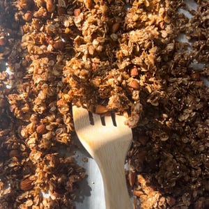 May include: A close-up of a baking tray filled with golden brown granola with almonds. A wooden spatula is used to stir the granola.
