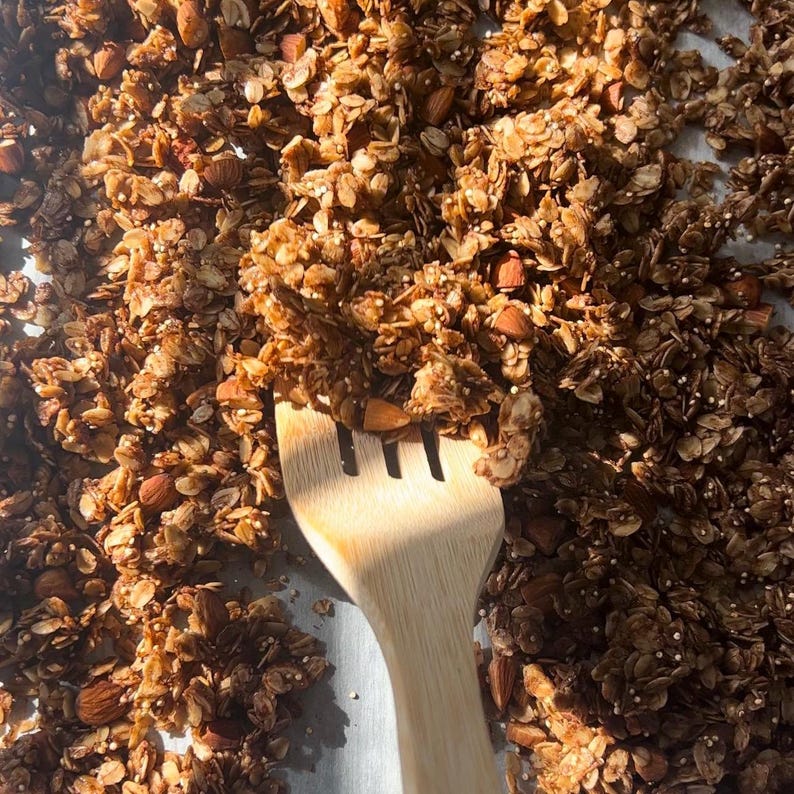 May include: A close-up of a baking tray filled with golden brown granola with almonds. A wooden spatula is used to stir the granola.