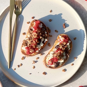 Puede incluir: Dos rebanadas de pan tostado cubiertas con queso blanco, mermelada roja y granola, presentadas en un plato blanco moteado. Un tenedor y un cuchillo dorados est&aacute;n colocados a la izquierda. Trozos de granola dispersos a&ntilde;aden inter&eacute;s visual.