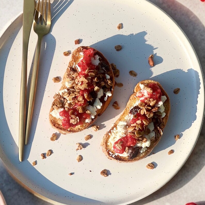 Puede incluir: Dos rebanadas de pan tostado cubiertas con queso blanco, mermelada roja y granola, presentadas en un plato blanco moteado. Un tenedor y un cuchillo dorados est&aacute;n colocados a la izquierda. Trozos de granola dispersos a&ntilde;aden inter&eacute;s visual.