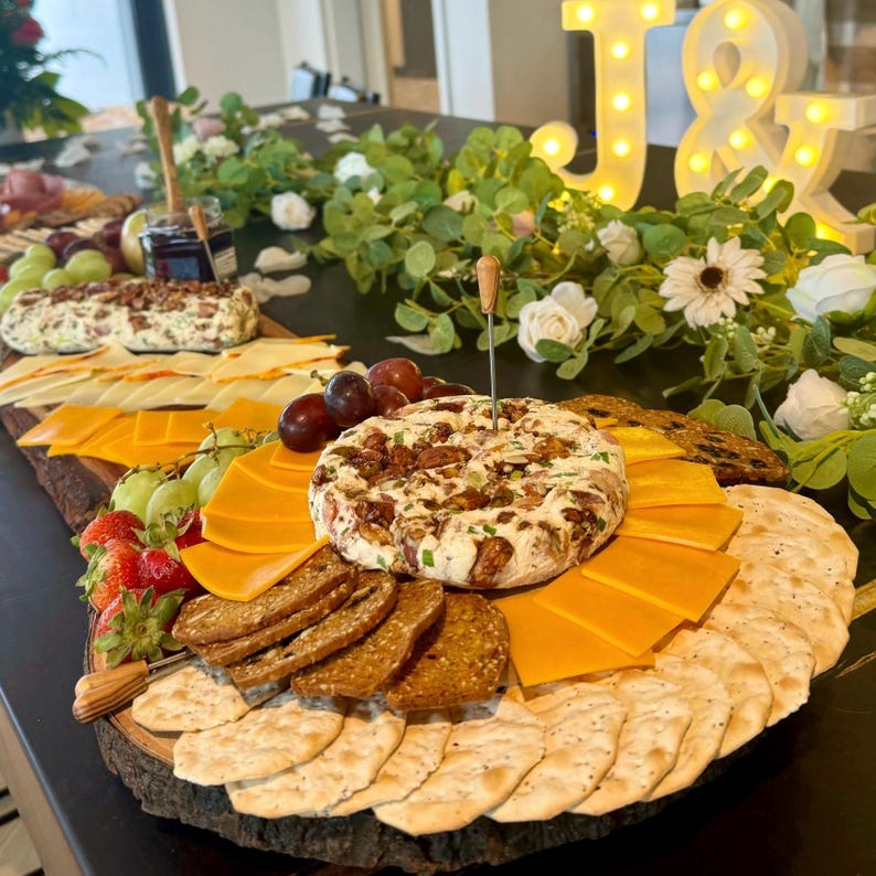 K&ouml;nnte beinhalten: Ein Holz-Servierbrett mit einer Auswahl an Snacks, darunter Cracker, K&auml;se, Obst und ein gro&szlig;er Dip mit N&uuml;ssen und Kr&auml;utern. Das Brett ist mit Gr&uuml;npflanzen und Blumen dekoriert.
