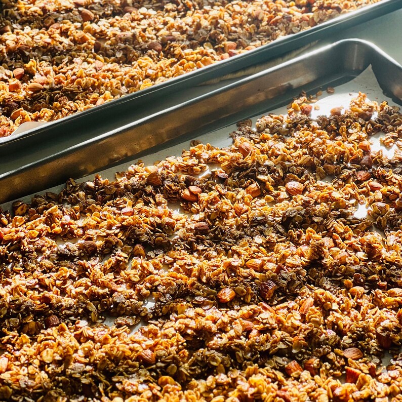 Puede incluir: Primer plano de dos bandejas de horno llenas de granola casera. La granola es una mezcla de avena, nueces y semillas de color marr&oacute;n dorado, con un aspecto ligeramente caramelizado. Las bandejas est&aacute;n forradas con papel de hornear.