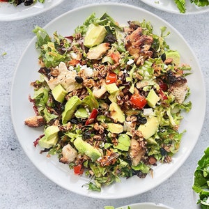 Puede incluir: Una ensalada vibrante en un plato blanco, con hojas verdes frescas, rodajas de aguacate, tomates en cubitos y pollo a la parrilla. La ensalada est&aacute; adornada con queso desmenuzado, nueces y bayas secas.