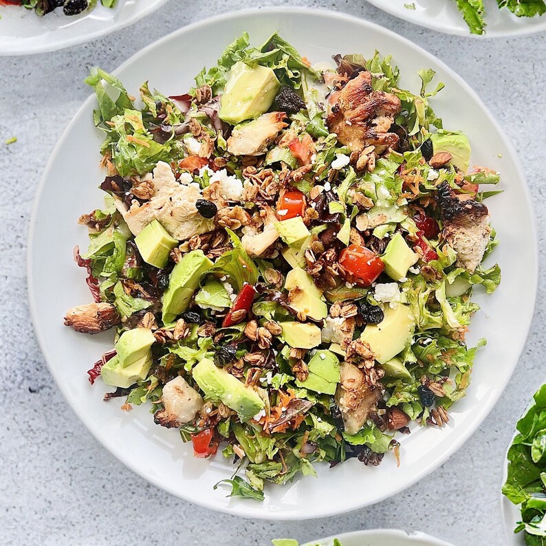 Puede incluir: Una ensalada vibrante en un plato blanco, con hojas verdes frescas, rodajas de aguacate, tomates en cubitos y pollo a la parrilla. La ensalada est&aacute; adornada con queso desmenuzado, nueces y bayas secas.