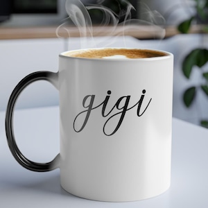 May include: White ceramic coffee mug with a black handle. The mug has the word "gigi" written in black cursive on the front.