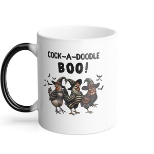 May include: White ceramic coffee mug with a black handle. The mug features a black and white image of three chickens wearing witch hats and the text "Cock-a-doodle Boo!" printed on it.