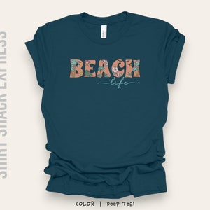 May include: A teal blue t-shirt with the text "Beach life" in a floral design. The text is in a peachy pink color with green leaves.
