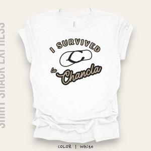 以下が含まれることがあります： 白地に「I survived la Chancla」という黒と茶色の文字が書かれたTシャツ。文字は茶色のフリップフロップの輪郭で囲まれています。