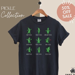 以下が含まれることがあります： 白地に緑色のピクルスのキャラクターが描かれた黒いTシャツ。キャラクターの名前は「パーティーピクルス」「スニーキーピクルス」「アロガントピクルス」など。Tシャツの上部には「PICKLE Collection」の文字、右上には円の中に「50% OFF SALE」の文字が書かれている。